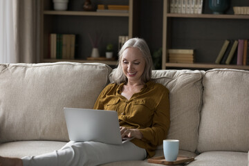Cheerful senior 50s laptop user woman enjoying leisure at home, resting on sofa in living room,...