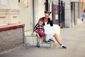 Cute european young mother with her son playing the red ukulele on the porch on the sidewalk. Walking around the city, learning to play musical instruments