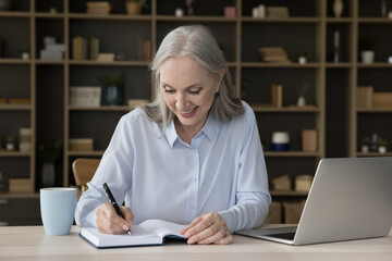 Happy busy senior freelance employee woman writing notes in note book at laptop. Senior student...