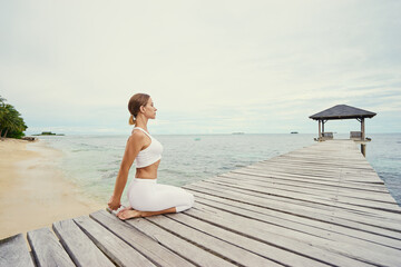 Fototapeta na wymiar Yoga on the beach. Young woman stretching on wooden pier with sea view.
