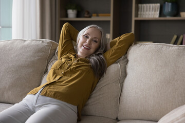 Happy pretty grey haired senior woman relaxing at home head shot portrait. Cheerful mid adult lady resting on comfortable couch, stretching body, leaning back, looking at camera, smiling