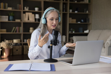 Elder grey haired teacher woman using laptop, wireless headphones, professional microphone, giving...