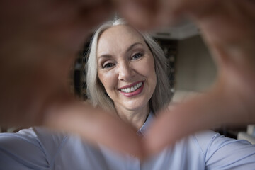 Happy grateful grey haired lady looking at camera through hand finger heart shaped frame. Pretty retired mature woman making gesture of love, empathy, tenderness. Close up portrait