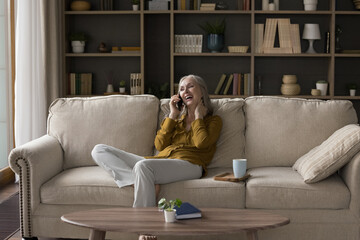 Cheerful excited mature smartphone user woman talking on cell, enjoying phone call conversation, resting on couch at cup of hot drink, speaking, laughing. Communication, retirement concept