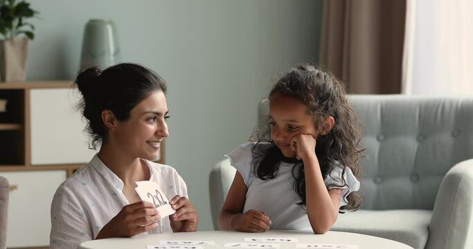 Smiling Indian mother nanny study multiplication tables with little junior schoolgirl show cards with printed sums ask questions praise for correct answers. Mommy teach math little kid in funny game