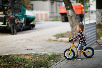 Fototapeta na wymiar cute european boy on balance bike watches how the road repair equipment works in the city, boys and construction equipment, boy looking at the skating rink