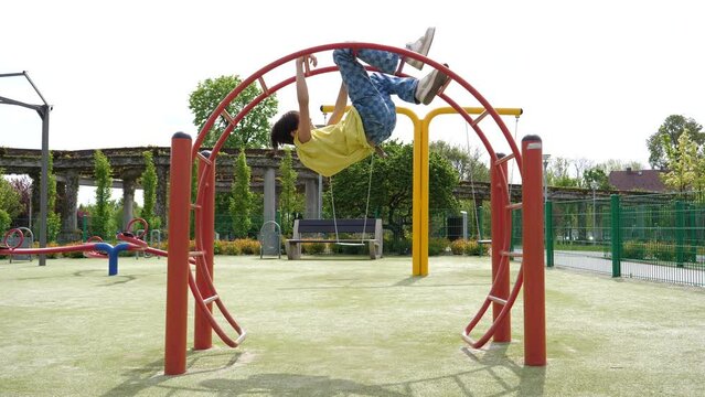 Teenager kid boy alone on playground for children hang upside down on a bar