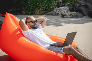 Young man working on his laptop on the beach while sitting on an inflatable sofa