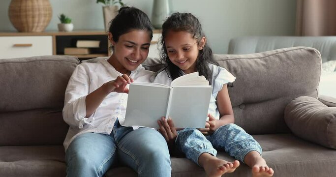 Young Indian mother sit on sofa engaged in reading interesting story with little daughter preschooler. Happy smiling mom small child girl turn printed family album photo book pages watch pictures talk