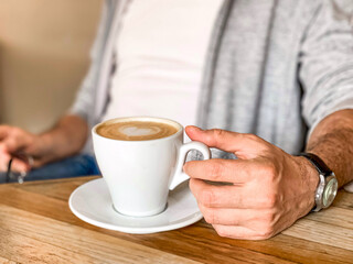 Young man is drinking morning coffee in cafe white ceramic mug with saucer on wooden table. Man in white t-shirt and grey sportive jacket is drinking his morning coffee in a cafe shop. Male hand touch