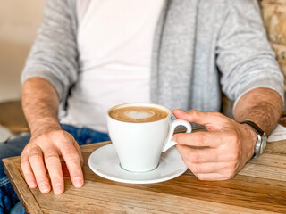 Young man is drinking morning coffee in cafe white ceramic mug with saucer on wooden table. Man in white t-shirt and grey sportive jacket is drinking his morning coffee in a cafe shop. Male hand touch