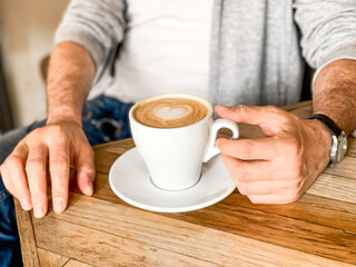 Young man is drinking morning coffee in cafe white ceramic mug with saucer on wooden table. Man in white t-shirt and grey sportive jacket is drinking his morning coffee in a cafe shop. Male hand touch