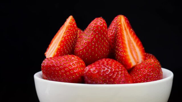 Strawberries without tails lie in white bowl or plate. It rotates on its axis on black background. Strawberry dessert. Appetizing pieces of incised berries. Vitamins and luxury.