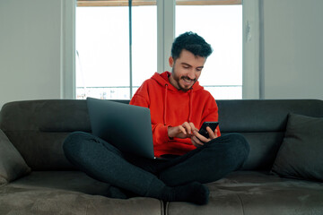 The young man sitting on the sofa has a laptop on his lap and is surfing the internet with his phone, young adult having a relaxed and enjoyable moment