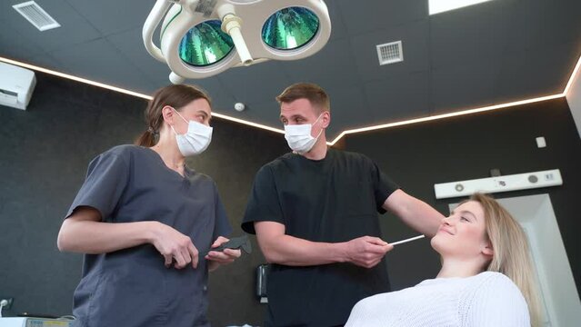 A plastic surgeon consults a young attractive woman. The doctor uses a pointer to show the main points of treatment on the face.He is assisted by a nurse