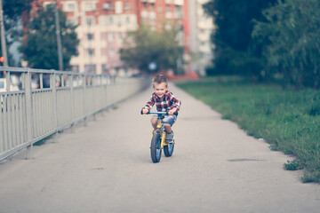 cute blond european boy rides balance bike on the road in city park, child rides in city in summer, lifestyle and summer vacation in town