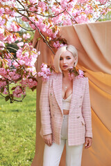 Fototapeta premium Gorgeous woman with short white hair on a beige background in the park near the sakura, which bloomed in spring with pink flowers.
