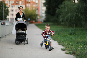 A mother with a stroller with a baby and a son on a balance bike walks around the city, a mother...