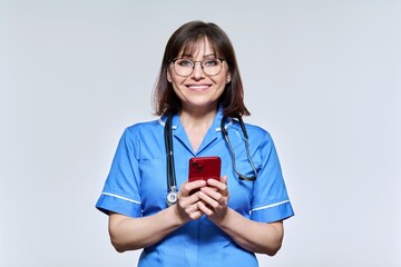 Portrait of female nurse with smartphone in hands, looking at camera on light background