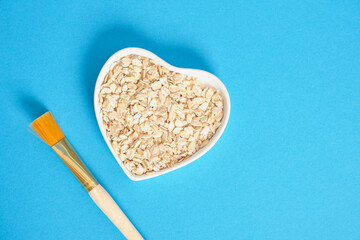 cosmetic brush and oatmeal in heart shaped plate on blue background, homemade face and hair mask
