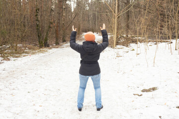 Young woman in jeans, black jacket and orange hat raised her hands up to God. Early spring in the forest. Back view