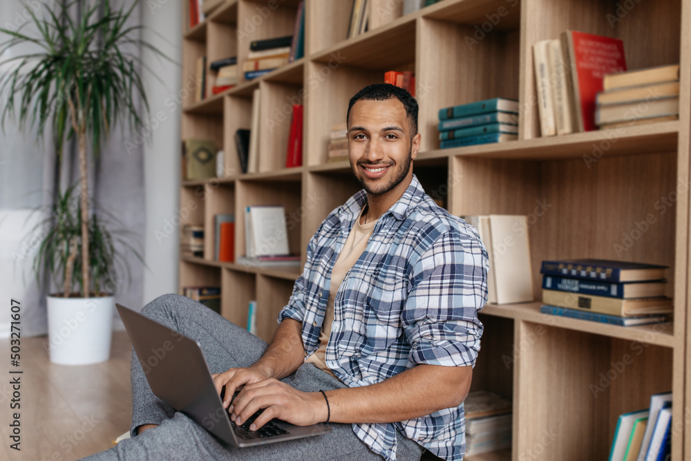 Poster portrait of happy arab man sitting with laptop and leaning on bookshelf, working on pc laptop and sm