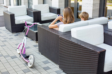 Children sit on the street, looking at the smartphone screen. Little school children sitting at a table on the veranda of a cafe, using a mobile phone. modern technologies, gadgets, entertainment.