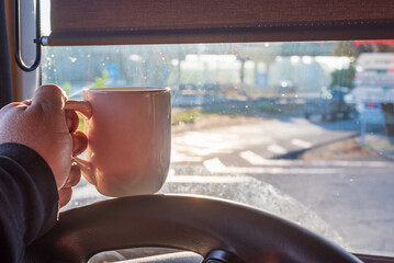 Hand holding a cup of coffee on the steering wheel of a truck. Truck driver in a moment of rest in...