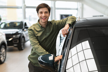 Fototapeta na wymiar Cheerful young man posing near new automobile after purchase, buying modern car at dealership