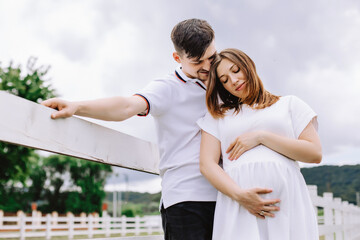Naklejka premium A man hugs his pregnant wife near the white fence on a background of cloudy sky