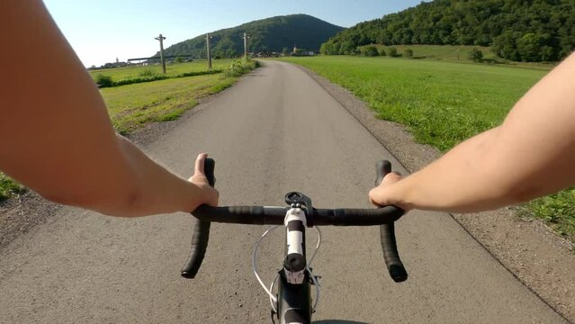 Caucasian female road cycling racer riding on an empty narrow country lane, watching a green countryside and mountain scenery in front, point of view shot.
