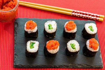 Sushi pieces on a square slate plate, accompanied by red caviar substitute, on a red background.