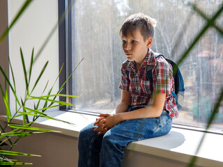 A portrait of sad boy with backpack sitting on the window sill frustrated sad expression, teeenager filling depressed and unhappy, tired of school, waiting for summer holiday