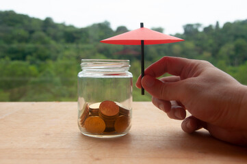 Hand holding small red umbrella over gold coins inside a jar on wooden table. Financial safety and investment concept.