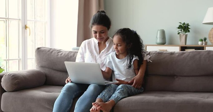 Young Indian mum nanny sit on sofa embrace adorable little daughter explain easy interesting learning material using pc. Caring mom teach small child to work in funny education app on laptop computer