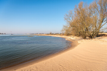 Sandy beach on the bank of the Dutch river Waal. It is a sunny day with a clear blue sky at the beginning of the spring season. River groynes made of basalt blocks are visible in the background.