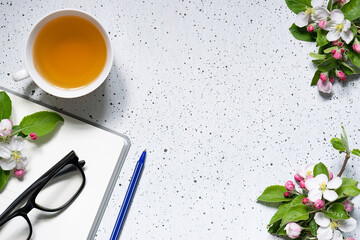 Artistic composition with blank notepad, glasses, cup of herbal tea and beautiful blooming apple twigs on white marble background with copy space for your design. Festive office desktop concept.