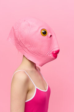 A woman in a pink fish head mask stands in profile against a pink background and looks into the camera with one yellow eye, a crazy conceptual Halloween costume