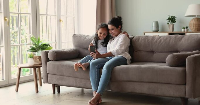 Loving Indian mother little daughter preschool age child engaged in online activity use smartphone resting on sofa at living room. Young nanny watch funny movie on cell together with small gen z girl
