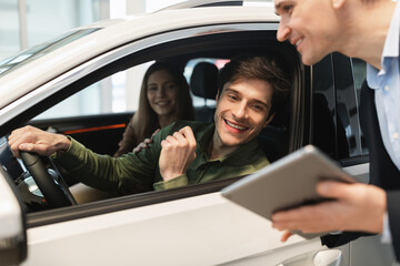 Young Caucasian couple clarifying car purchase details with salesman, looking at tablet screen in...