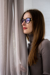 Young woman in glasses in the room looks out the window