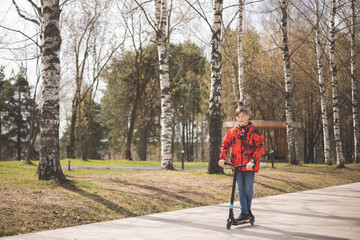 Happy teen riding scooter in park. Ecological urban transport. Caucasian teenager boy in red jacket having fun driving scooter through urban city street on spring day. Child walking outdoors.