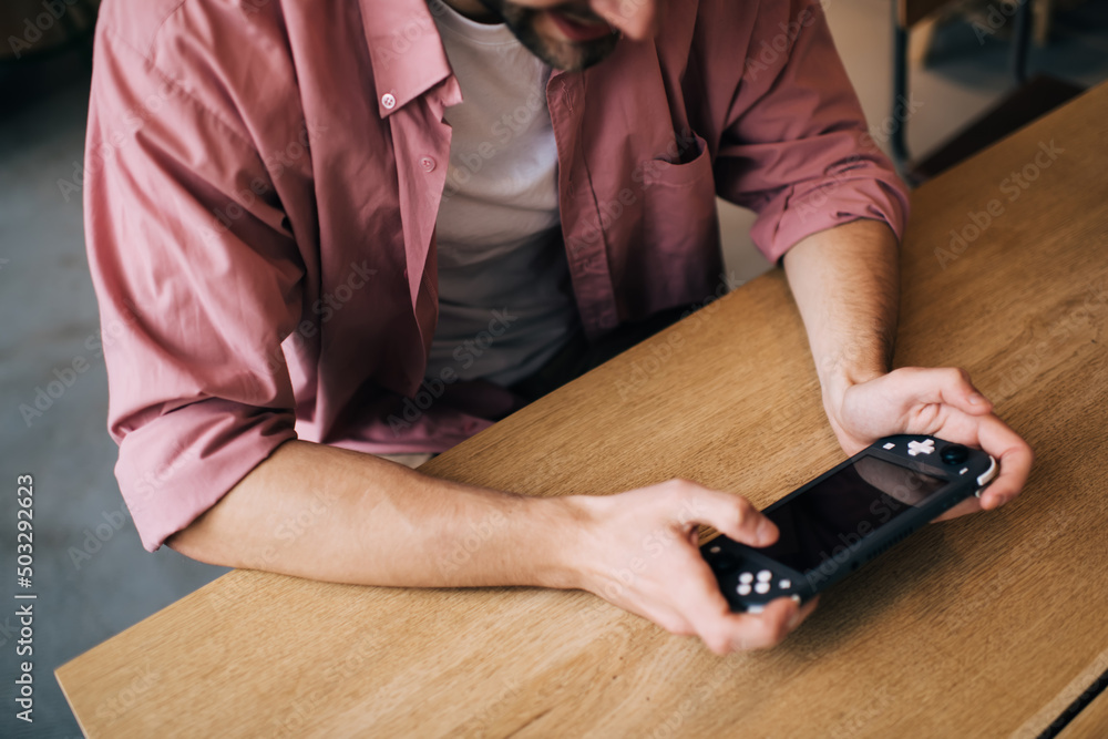 Poster cropped image of millennial male holding gamepad controller for playing video game indoors, unrecogn