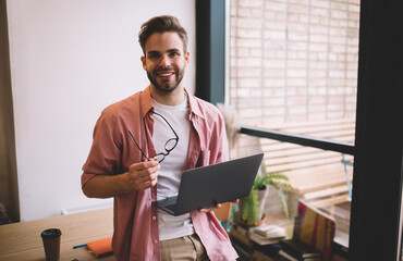 Half length of happy male freelancer with laptop device and spectacles in hands smiling at camera...
