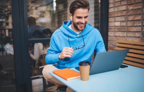 Cheerful male freelancer browsing funny video content while networking in social media using netbook technology in street cafe, happy hipster guy enjoying free time for online blogging via laptop