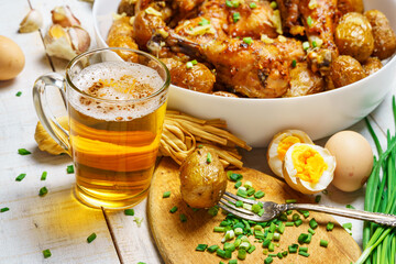 lunch in rural style, tasty food - glass of beer, fried chicken meat and potatoes, garlic and green onions, cheese, boiled eggs and salt, cooked food on a white wooden boards