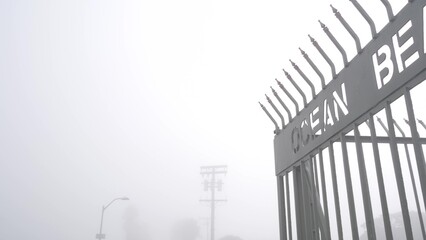 Metal gate on Ocean Beach pier in fog, misty California coast, USA. Foggy moody cloudy weather on San Diego shore. Calm tranquil atmosphere. Waterfront boardwalk entrance in gloomy depressive haze.