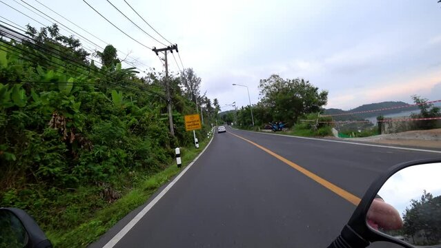 View of the road and the side mirror of the motorcycle. Clean smooth road in Asia. Cars and scooters are passing by. A guy in a helmet on a bike. Trees and palm trees grow along the road. Phuket, Thai