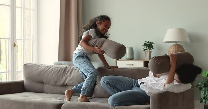 Grownup elder sister play with younger girl child on cozy couch imitate funny battle act shooting using sofa cushions as guns. Laughing Indian mom enjoy imaginary fight on pillows with little daughter