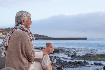 Attractive relaxed caucasian senior woman sitting on the pebble beach at sunset light admiring...